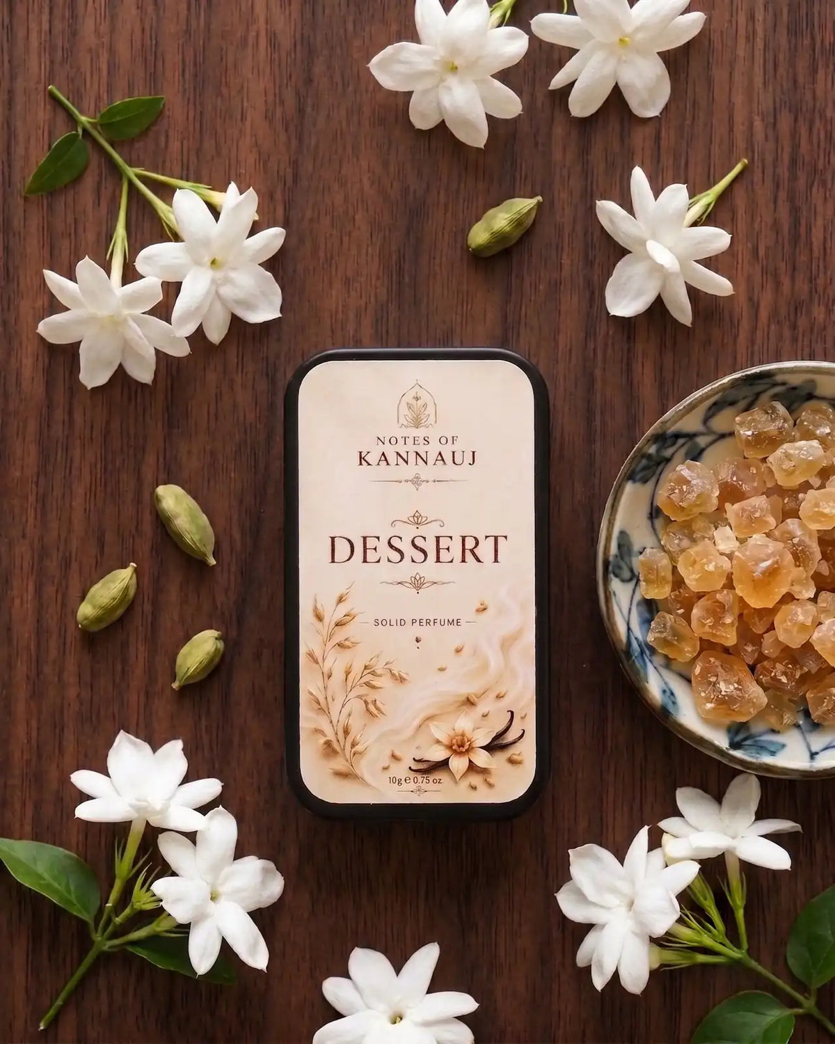 A rectangular perfume tin labeled 'Notes of Kannauj Dessert' is arranged on a dark wooden surface with white jasmine flowers, green leaves, and a small bowl of amber-colored crystals.