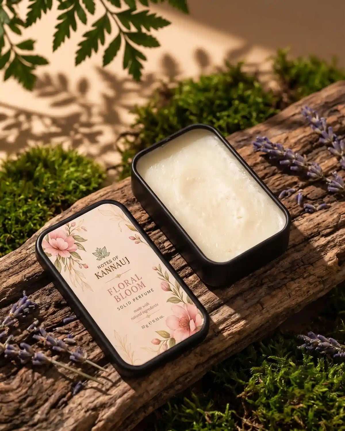 An open black metal tin of Notes Of Kannauj Floral Blossom solid perfume sits on a rustic log surrounded by green moss, dried lavender sprigs, and soft leaf shadows.